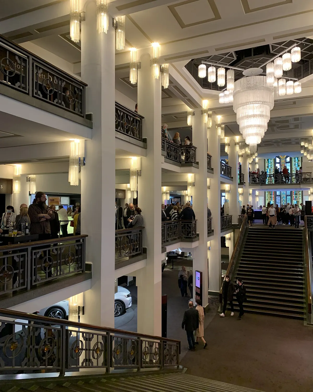 A broad staircase and tall white columns in the foyer of the Palast, with a large glass chandelier and balconies with ornate railings