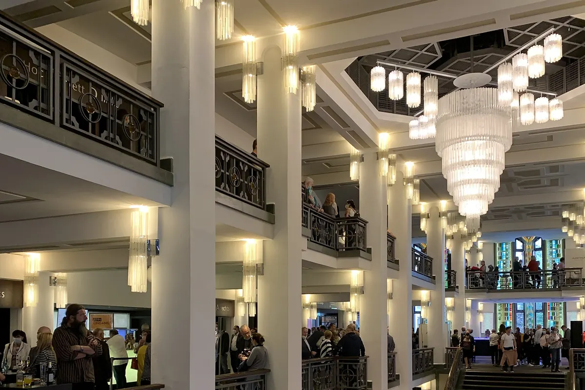 The foyer of a theatre with a wide staircase, tall white columns, a glass chandelier and balconies with ornate railings